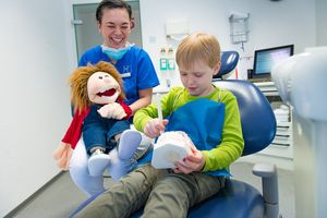 Eine Behandlerin mit einer Handpuppe sitzt neben einem Kind in einem Zahnarztstuhl, während das Kind das Zähneputzen an einem Zahnmodell übt.