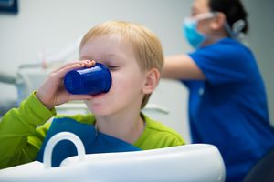 Ein kleiner Junge auf einem Zahnarztstuhl trinkt aus einem blauen Becher, während im Hintergrund eine Behandlerin mit Maske arbeitet.