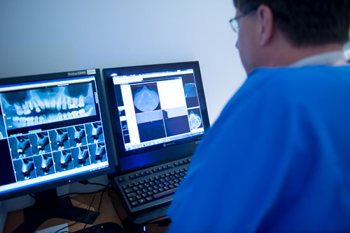 Eine Person im blauen Hemd prüft zahnärztliche Röntgenaufnahmen und medizinische Bilder auf zwei Computermonitoren in einer klinischen Umgebung.