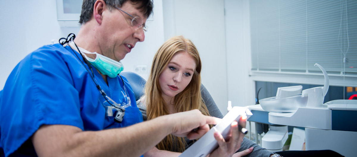 Ein Zahnarzt im Kittel erklärt einer sitzenden Patientin in einer Zahnarztpraxis Informationen auf einem Tablet.