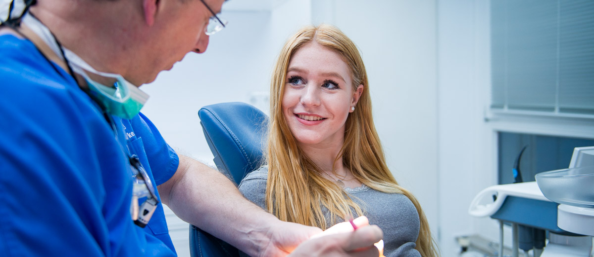 Ein Zahnarzt in blauem Kittel spricht mit einem lächelnden Patienten auf einem Zahnarztstuhl in einer modernen Klinik.