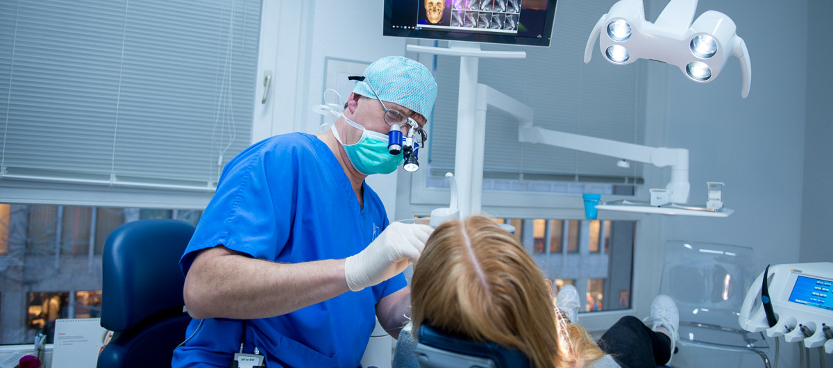 Ein Zahnarzt in blauem Kittel und Maske untersucht die Zähne eines Patienten in einer modernen Zahnklinik. Im Hintergrund sind zahnmedizinische Geräte und ein Monitor zu sehen.