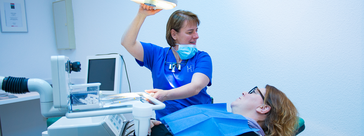 Eine Zahnärztin stellt eine Deckenleuchte ein, während ein Patient mit Schutzkleidung auf einem Zahnarztstuhl in einer Klinik liegt.